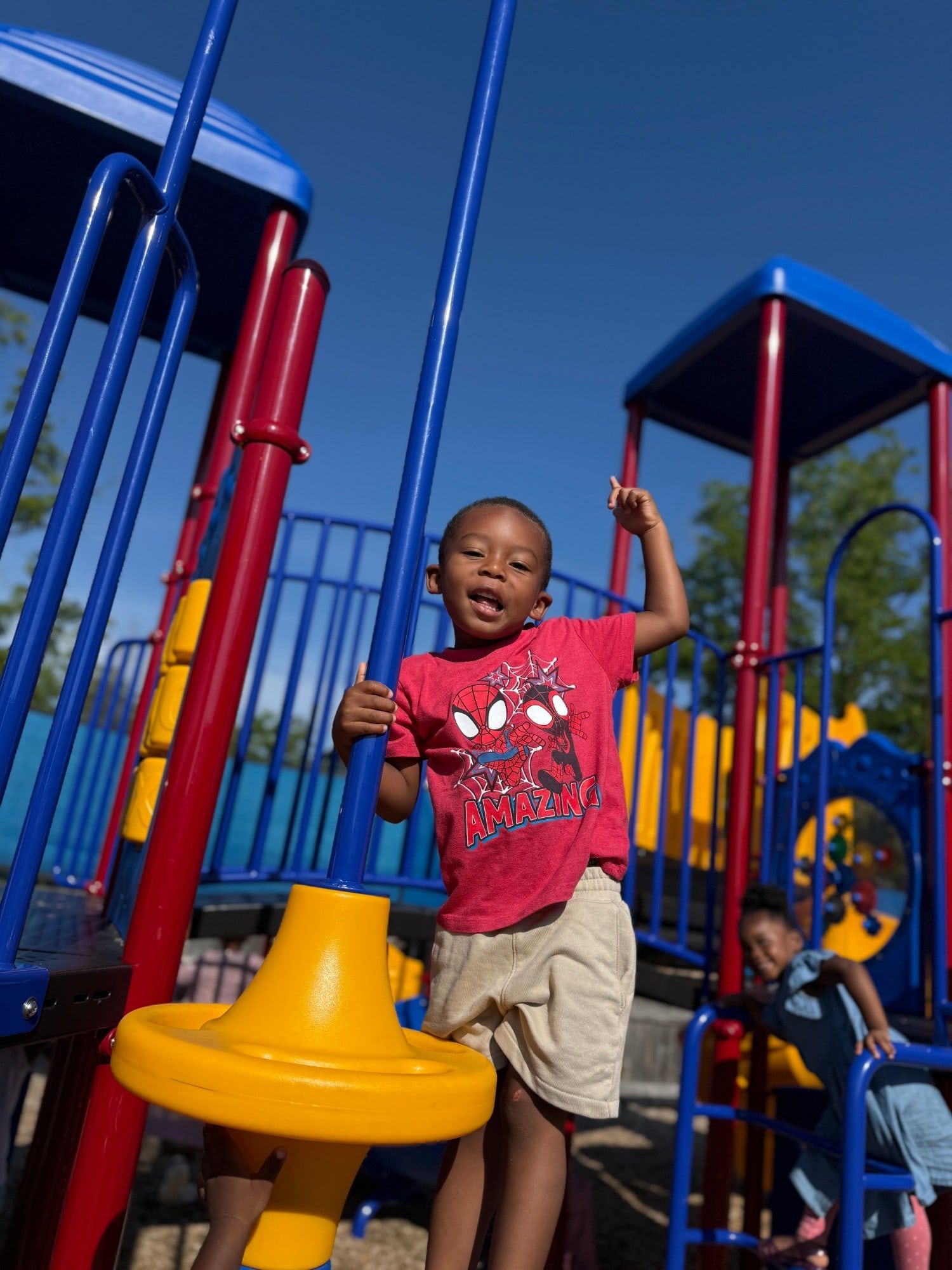 A boy in a red Spider-Man shirt cheering with one arm raised on the playground