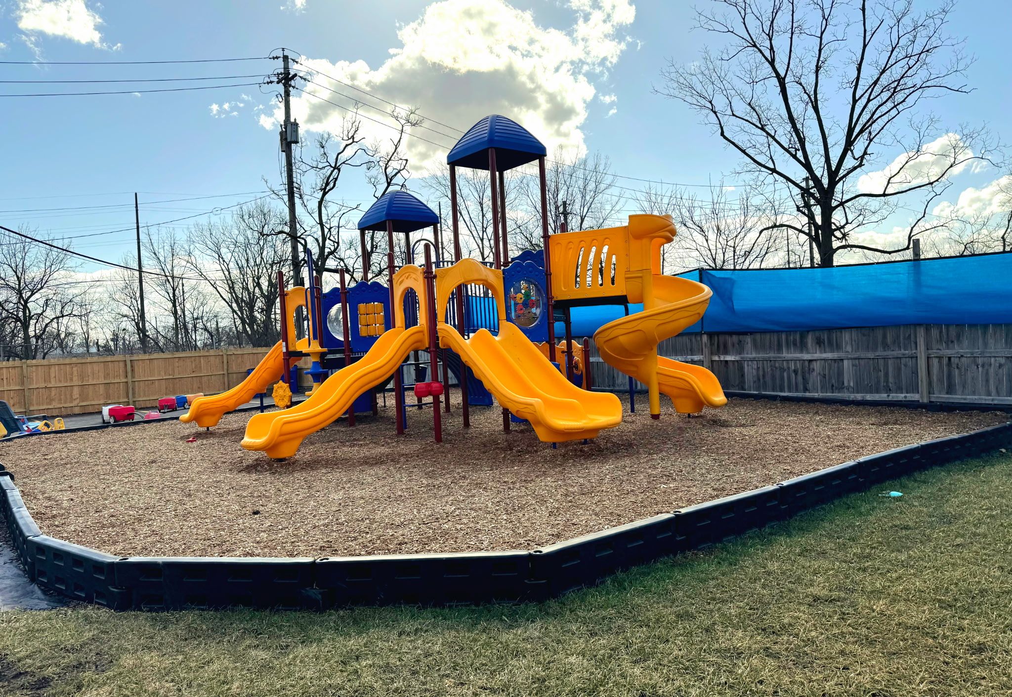 Our fenced playground: a yellow and blue climbing structure with three slides on a wood-chip surface